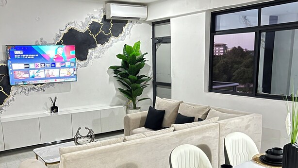 A modern living room with a sofa, dining table, chairs, a TV, and a decorative wall feature. - Modern 3BR Hideaway in Masaki | Pool, Gym & Prime Location (Dar es Salaam)