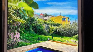 Una piscina con cubierta de madera, rodeada de vegetación y con vista a un edificio amarillo al fondo.