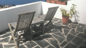 Una terraza con dos sillas de madera y una mesa, plantas en macetas y vista al mar.