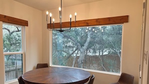 Comedor con una mesa redonda de madera, cuatro sillas, una araña y ventanas amplias con vista a árboles.