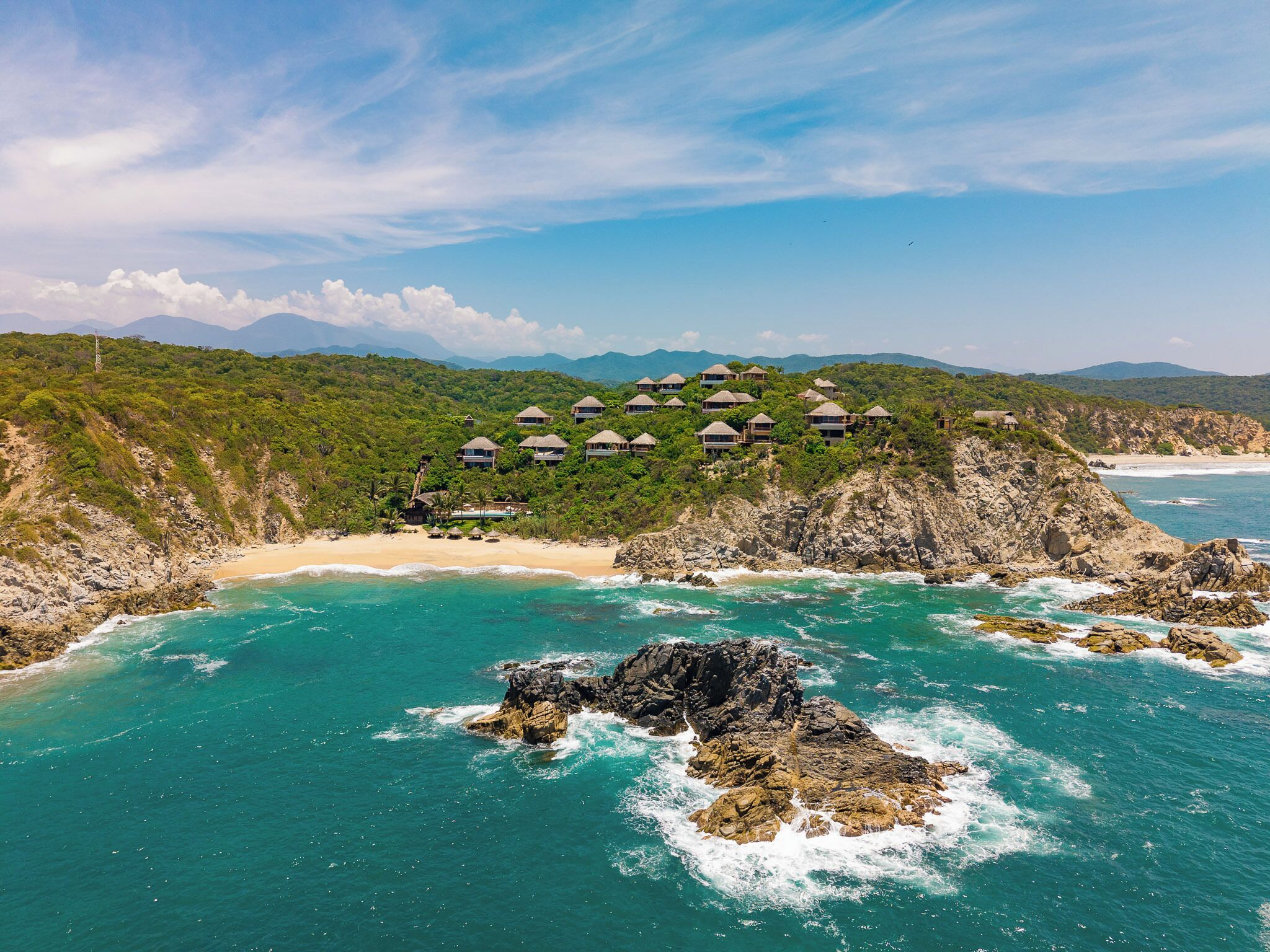 Une station balnéaire dotée de bungalows sur pilotis, situés sur un promontoire rocheux, d’une plage de sable et d’eaux turquoise.