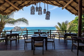 Een houten terras met tafels en stoelen met uitzicht op een strand met een rotsformatie en palmbomen.