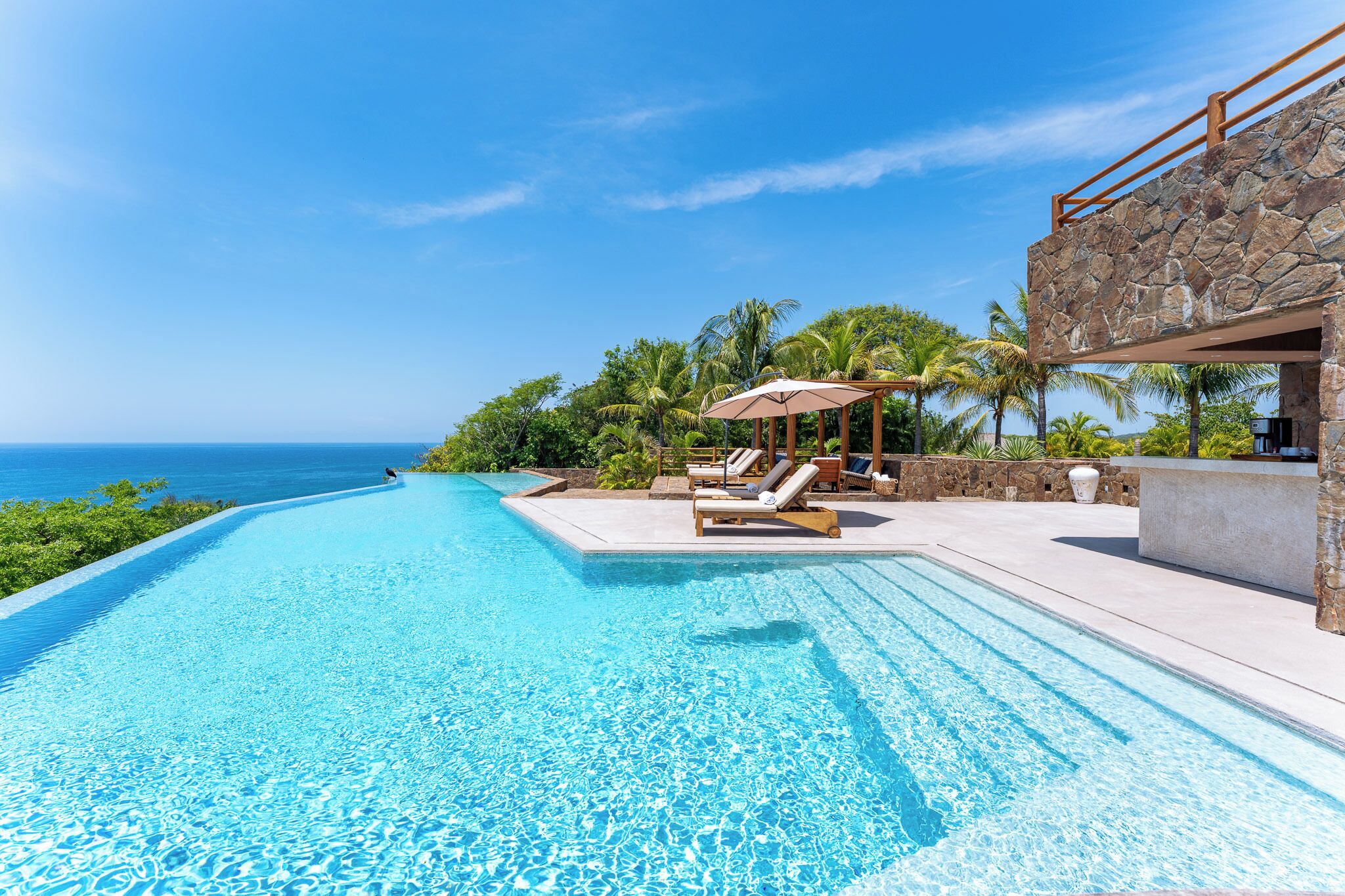 Une magnifique piscine à débordement d’un bleu profond, agrémentée de chaises longues et de parasols, et entourée d’une végétation luxuriante et d’un mur de pierre.