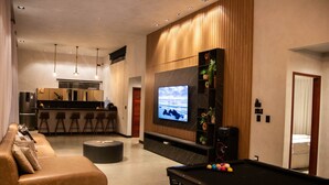 A modern living room with a pool table, a sofa, a TV mounted on a wooden wall, and a kitchen area in the background.