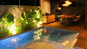 A swimming pool with a wooden deck, a stone wall, and a dining area with a table and chairs.