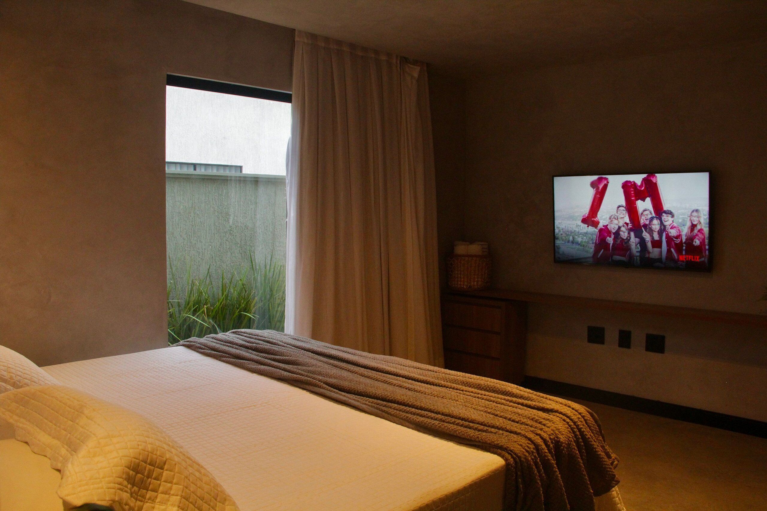 Ein Hotelzimmer mit Bett, Fernseher an der Wand, Fenster mit Vorhängen und Blick auf Grünfläche.