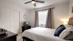 Une chambre à coucher avec un lit, un téléviseur posé sur un meuble, un ventilateur de plafond et une fenêtre avec des rideaux.