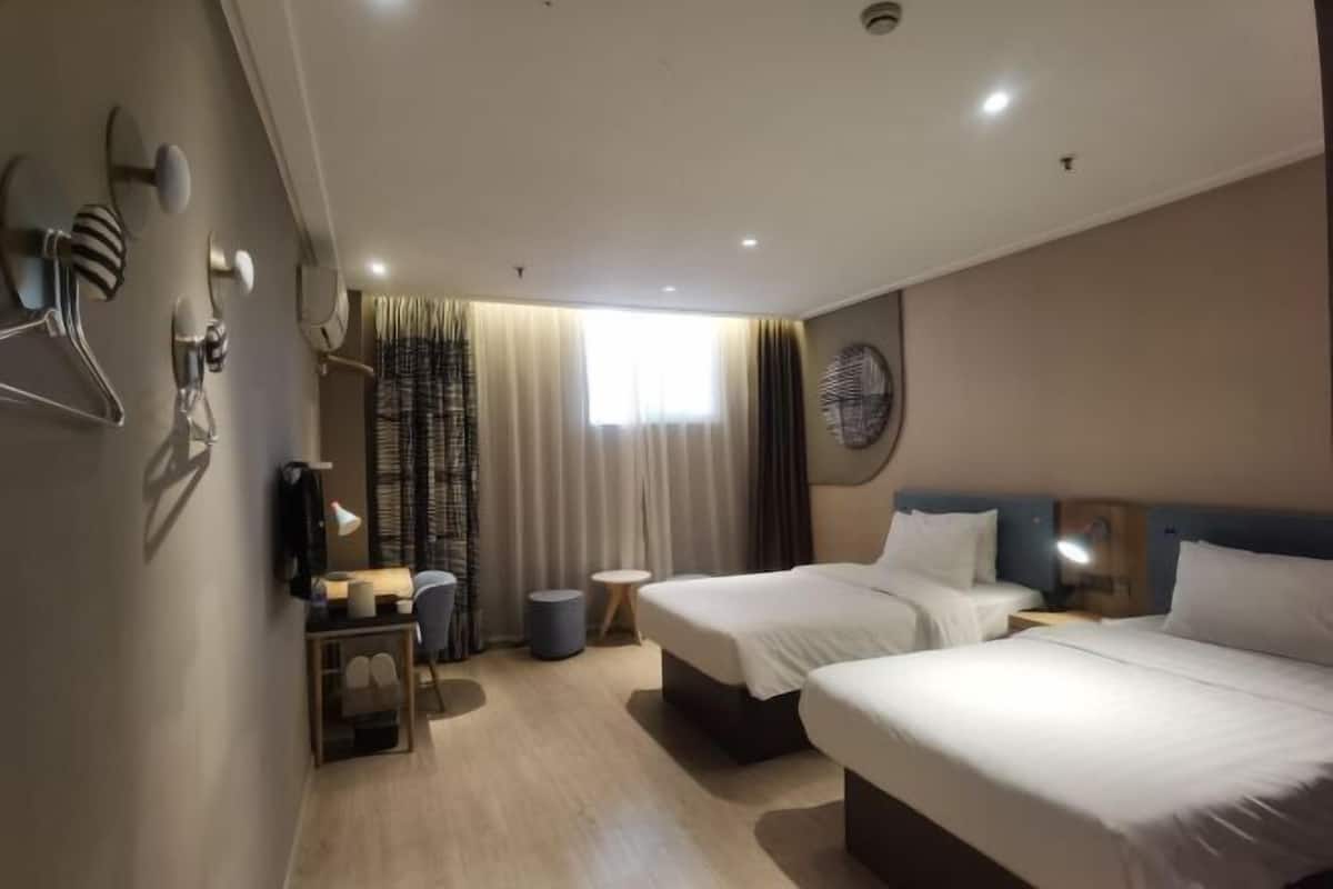Ein Hotelzimmer mit zwei Betten, einem Schreibtisch mit Stuhl, einem kleinen Tisch mit Lampe und einem Fenster mit Vorhängen.