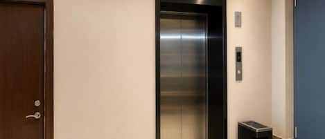 An elevator with a metallic finish, a door with a handle, and a trash bin beside it.