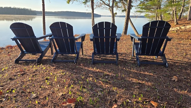 Three Adirondack chairs facing a calm lake with trees in the background.