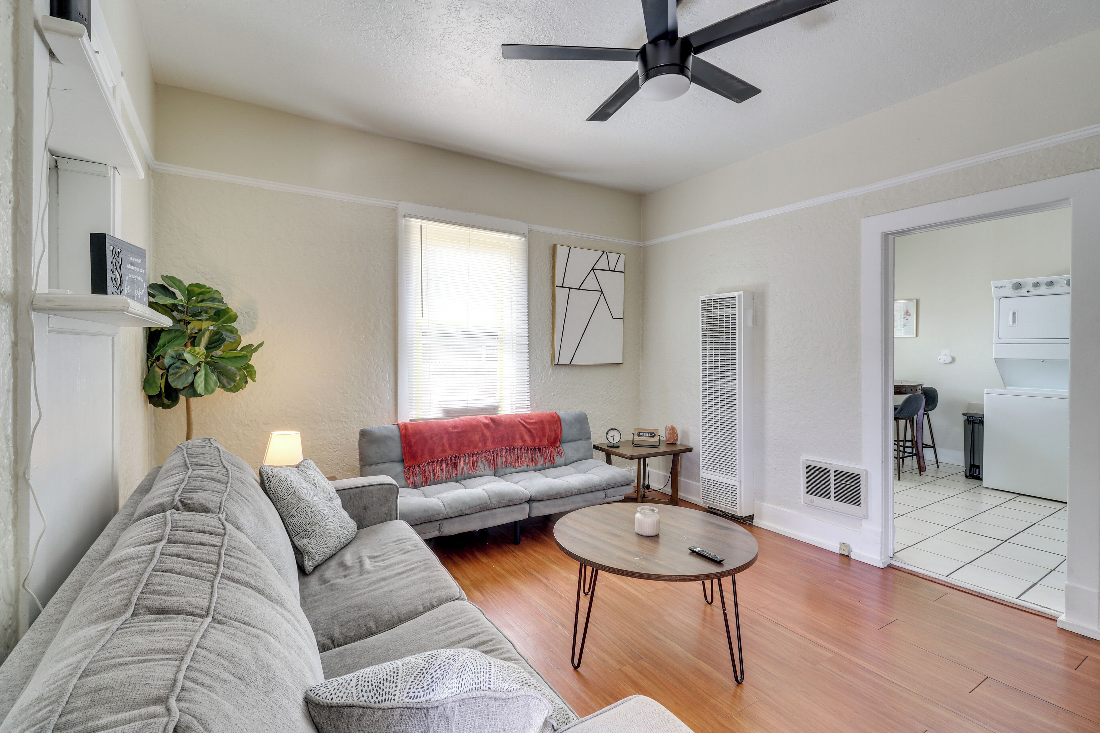 A living room with a grey sectional sofa, a round coffee table, a ceiling fan, a plant, and a view into the kitchen through an open door.