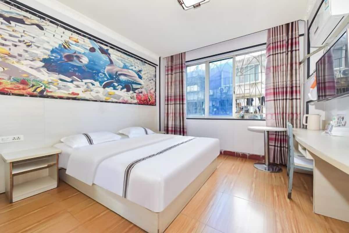 A hotel room with a bed, a small desk, a window with curtains, and a wall mural.