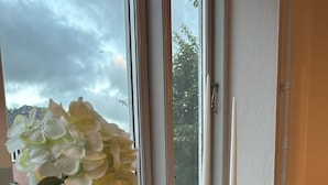 A clear view of a window with a vase of flowers on a white ledge, a tall candle, and a view of trees outside. - Kolding City Rooms (Kolding)
