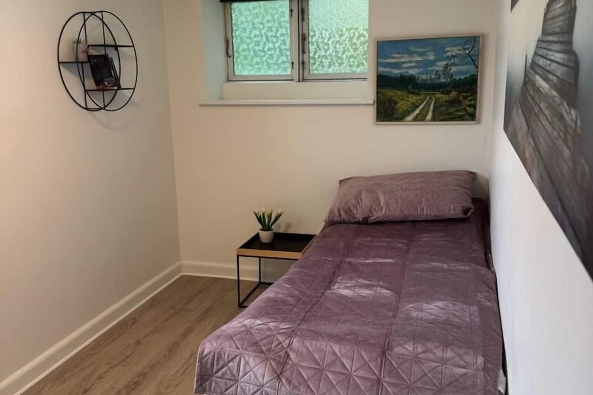 Une petite chambre avec un lit, une fenêtre, une étagère fixée au mur et un tableau.
