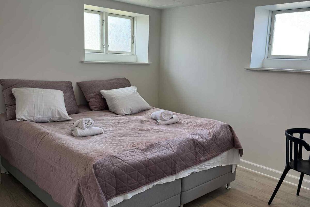 Un lit bien fait, recouvert d’une courtepointe matelassée, avec deux oreillers et une serviette enroulée. Il y a deux fenêtres à cadres blancs, une sur chaque mur, et un luminaire au plafond.