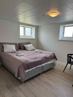 Ein ordentlich bezogenes Bett mit einer gesteppten Decke, zwei Kissen und einem zusammengerollten Handtuch. Zwei Fenster mit weißen Rahmen, eines an jeder Wand, und eine Deckenleuchte.