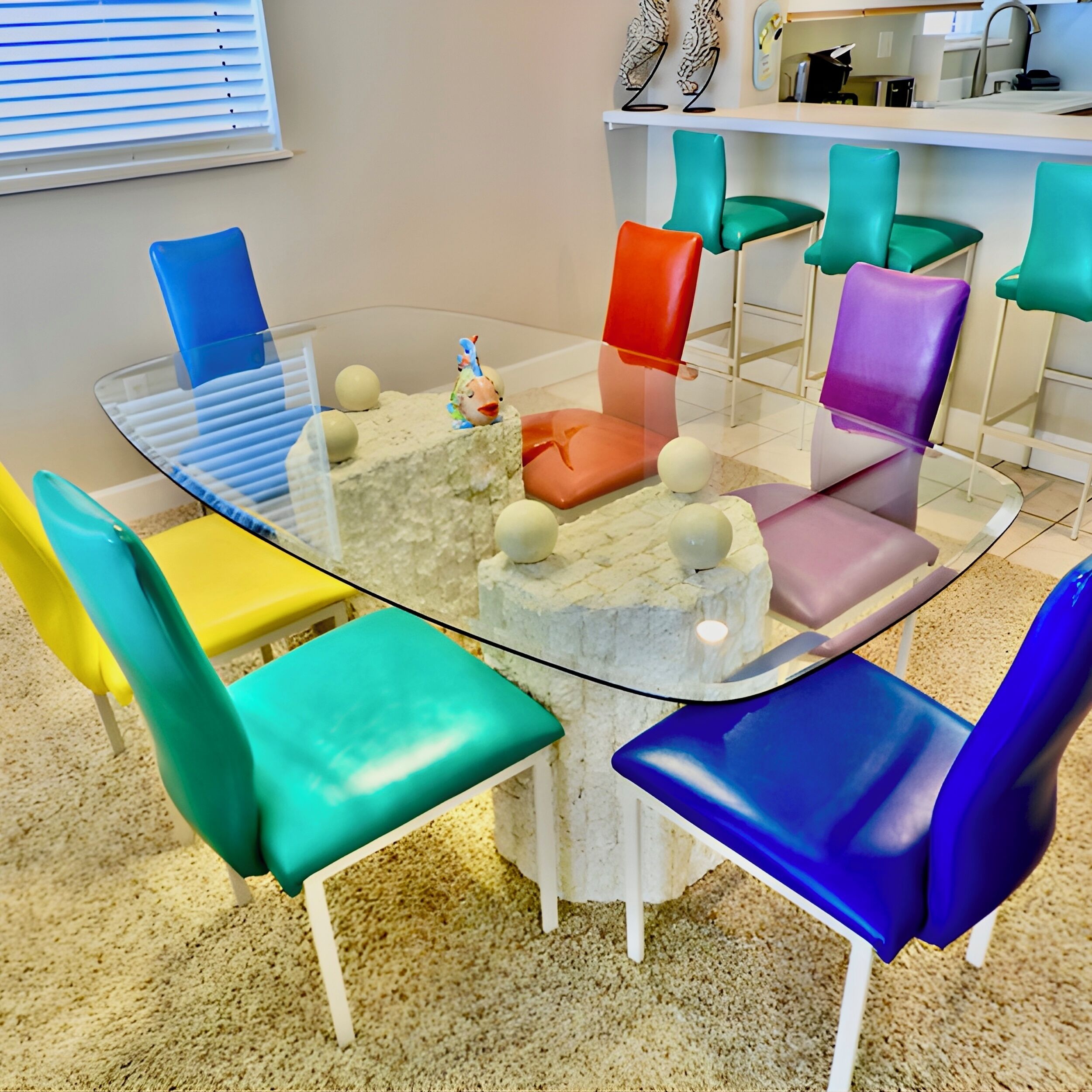 A round glass table surrounded by colorful chairs in a modern room with a white floor and a window with blinds.