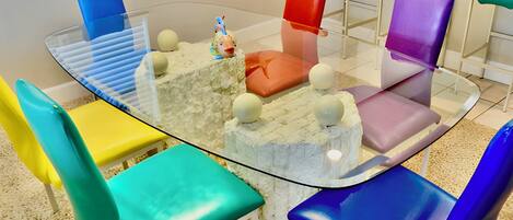 A round glass table surrounded by colorful chairs in a modern room with a white floor and a window with blinds.