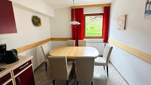 A dining area with a wooden table and four chairs, a coffee maker on a counter, and a window with red curtains. - Apartment in Fugen Near Ski Lift (Fuegen)