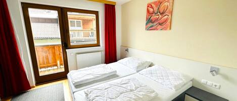 A hotel room with a bed, a small bedside table, a radiator, a window with a view of a balcony, and a painting on the wall.