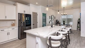 Una cocina moderna con armarios blancos, un refrigerador de acero inoxidable, una isla de mármol con taburetes altos, iluminación colgante y un comedor con mesa y sillas.