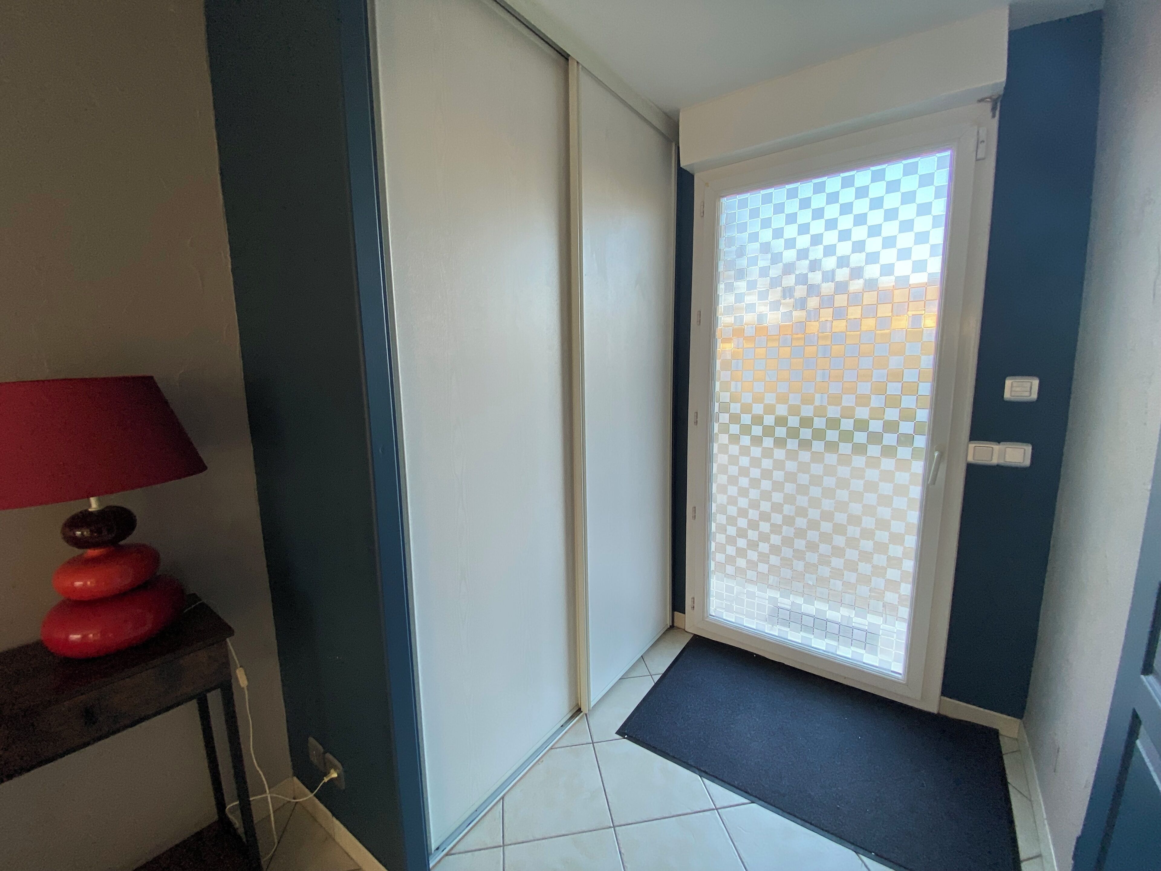 Un couloir avec une porte en verre à carreaux, une lampe rouge posée sur une table et un paillasson noir devant l’entrée.