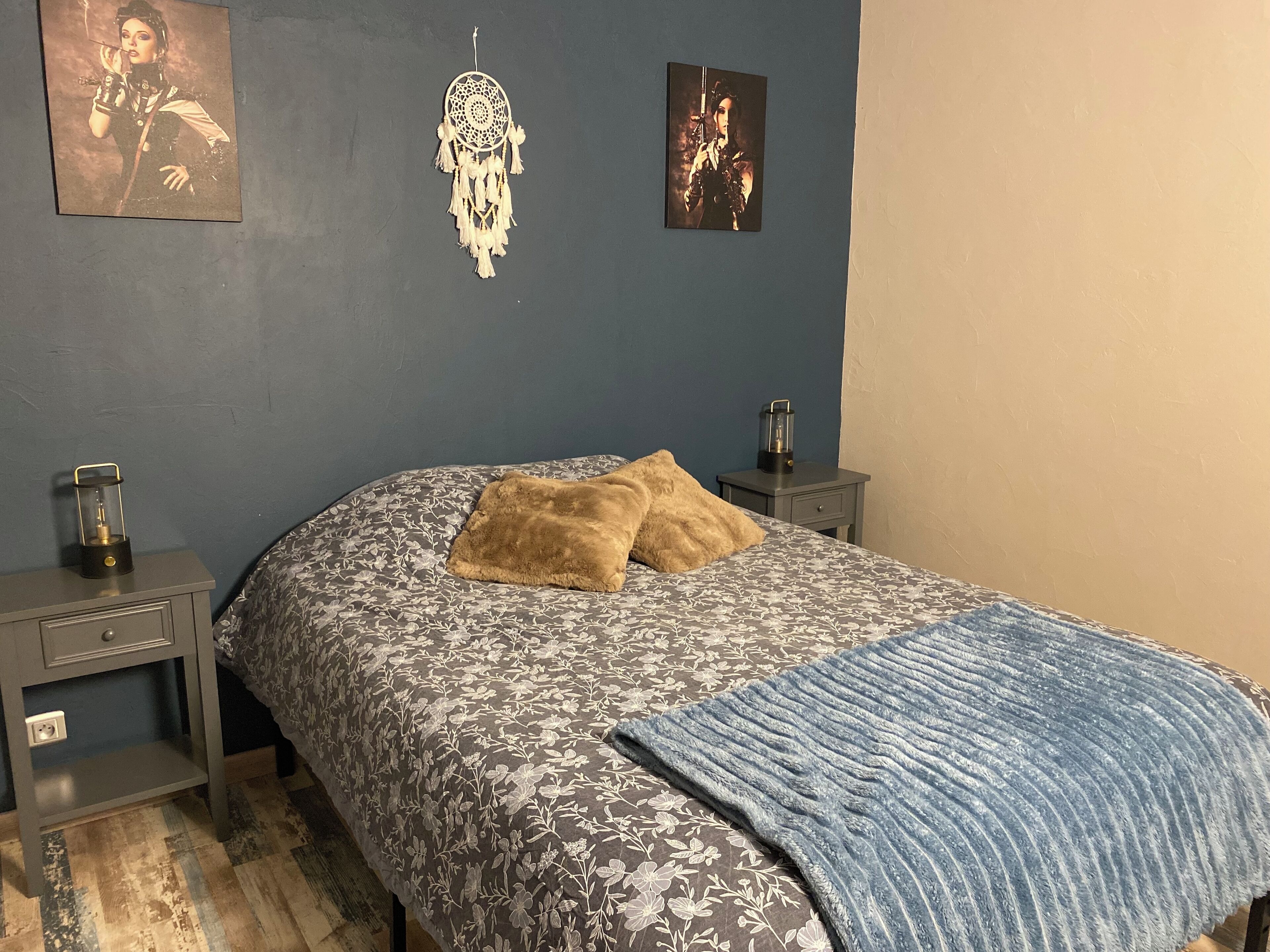 A bedroom with a floral-patterned bedspread, two pillows, a blue blanket, a dreamcatcher, and two wall-mounted pictures.
