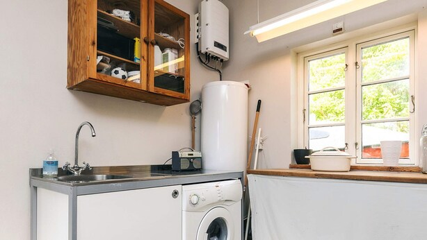 A compact kitchen with a washing machine, a wooden cabinet with various items, a window with a view of greenery, and a radiator.