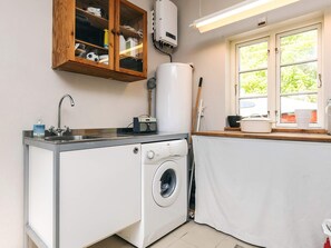 A compact kitchen with a washing machine, a wooden cabinet with various items, a window with a view of greenery, and a radiator.