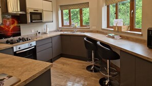 A modern kitchen with an island, bar stools, and large windows with views of greenery. - Luxury, secluded, Adult only Treehouse with hot tub, woodburner & stunning views (Glenshee)