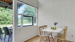 Et værelse med et stort vindue, et rundt bord med stole, og grøn vegetation udenfor.