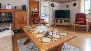 Een gezellige woonkamer met een houten plafond, een eettafel met een uniek patroon, een televisie, een rode fauteuil en een kast met glazen deuren.