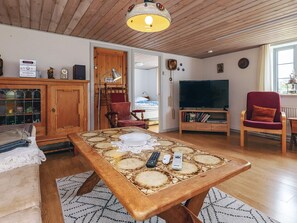 Ein gemütliches Wohnzimmer mit Holzbalkendecke, einem Esstisch mit weißen Tischdecken und einem roten Sessel sowie einem Fernseher.