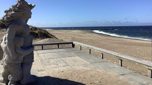 Een stenen standbeeld op een houten looppad dat naar een strand leidt, waar golven op de kust breken.