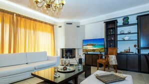 A modern living room with a sofa, coffee table, and TV. There is a bookshelf with decorative items and a view of the sea through the window. - Danae's House Elegant Residence in  Chalepa, Chania (Chania)