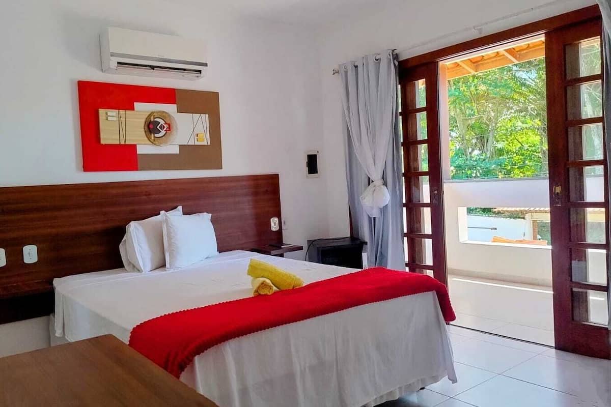 A hotel room with a wooden headboard, a bed with white bedding and a red accent, a small table, and a window with a view of greenery.