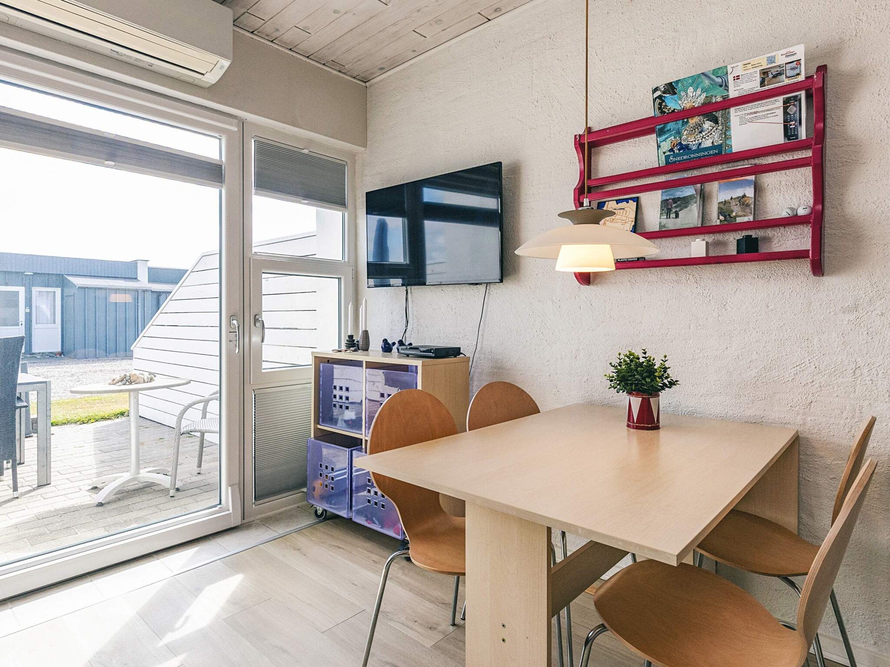 Una habitación moderna con una puerta corrediza de vidrio que da a un espacio exterior, un televisor de pantalla plana montado en la pared, una mesa de madera con sillas y un estante rojo con varios objetos.