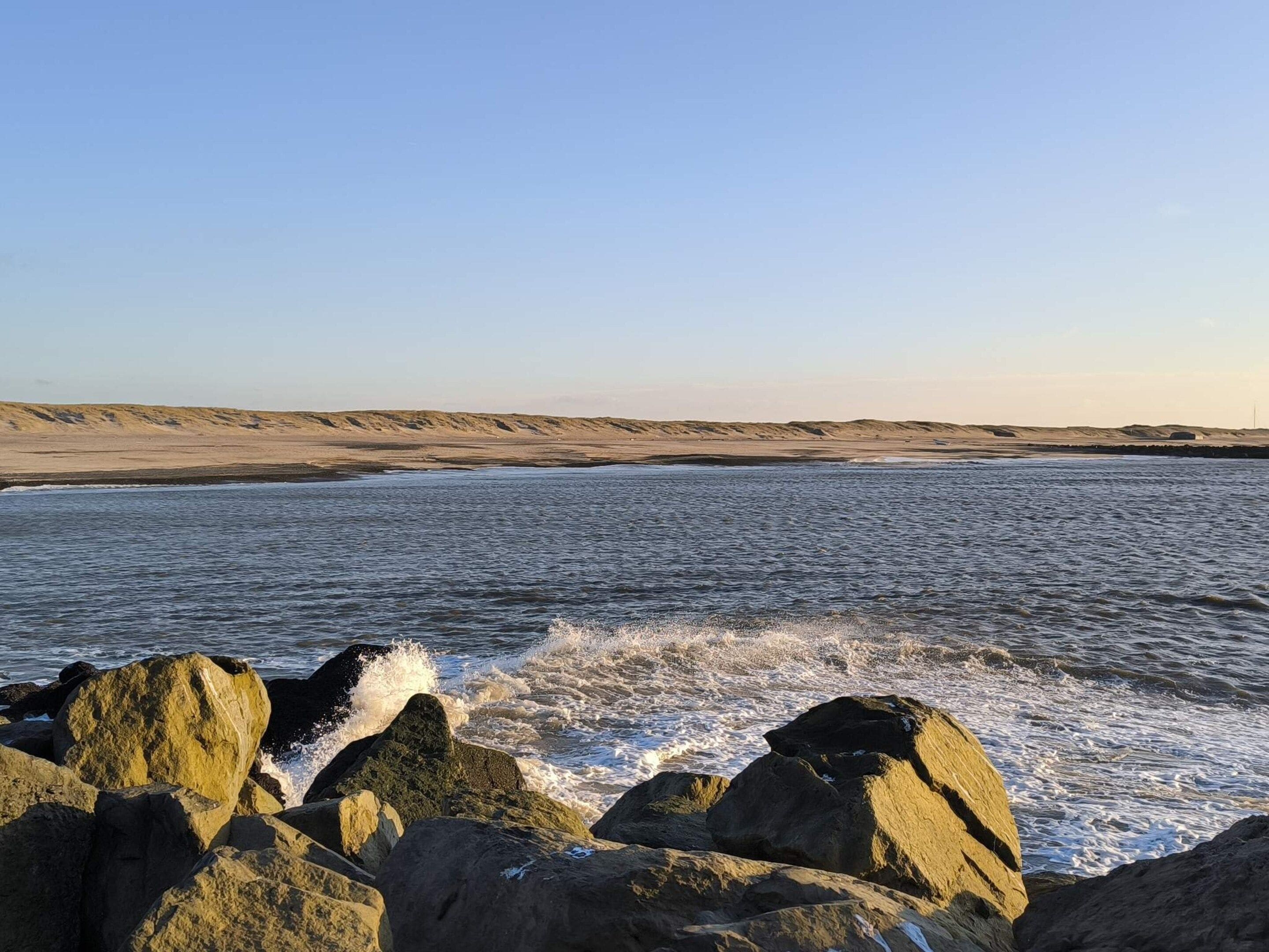 A coastal landscape with waves crashing against large rocks and a sandy beach in the background.