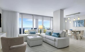 A modern living room with a large sofa, a small table, and a chair. There is a view of the ocean through large windows.
