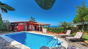 A swimming pool with lounge chairs, a red building, and a view of the sea.