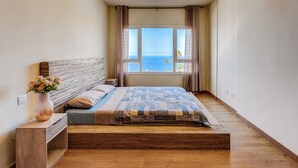 A bedroom with a large bed, a wooden headboard, a bedside table with a vase of flowers, and a window with a view of the sea.