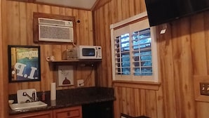 A compact kitchen with wooden walls, a microwave, a sink, and a window with blinds.