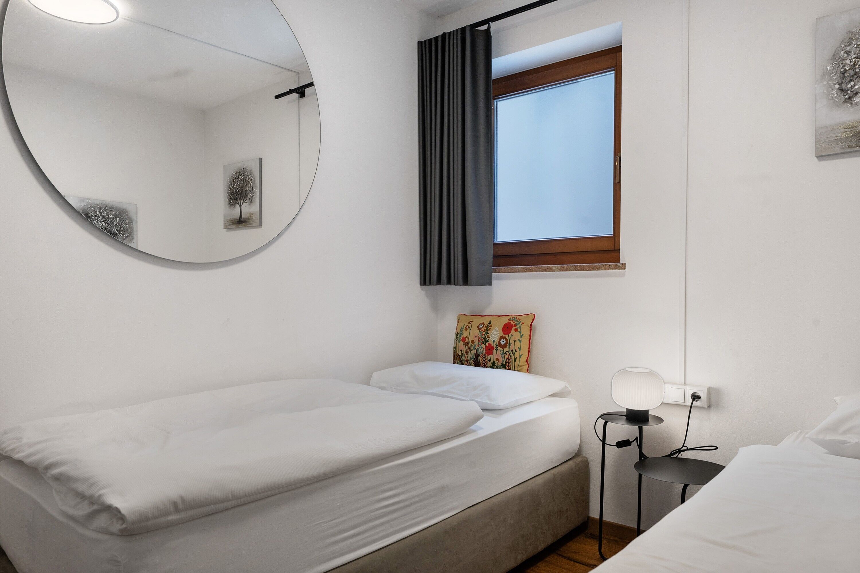 Un lit simple avec une tête de lit, une table de chevet avec une lampe, un miroir rond, une fenêtre avec des rideaux et un petit tableau décoratif mural.