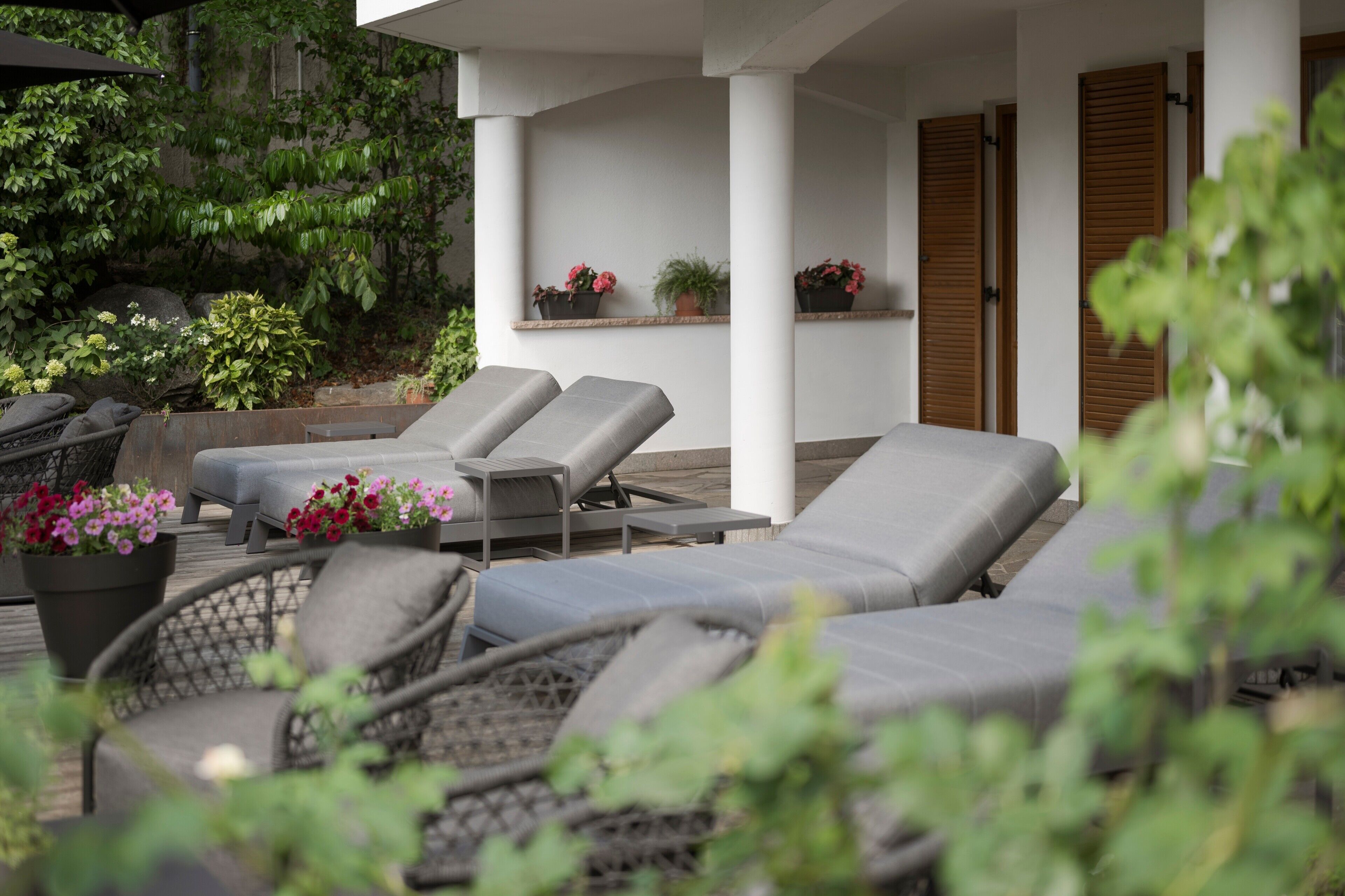 Een overdekt terras met lounge fauteuils en een rieten stoel, omgeven door potplanten en een houten terras.