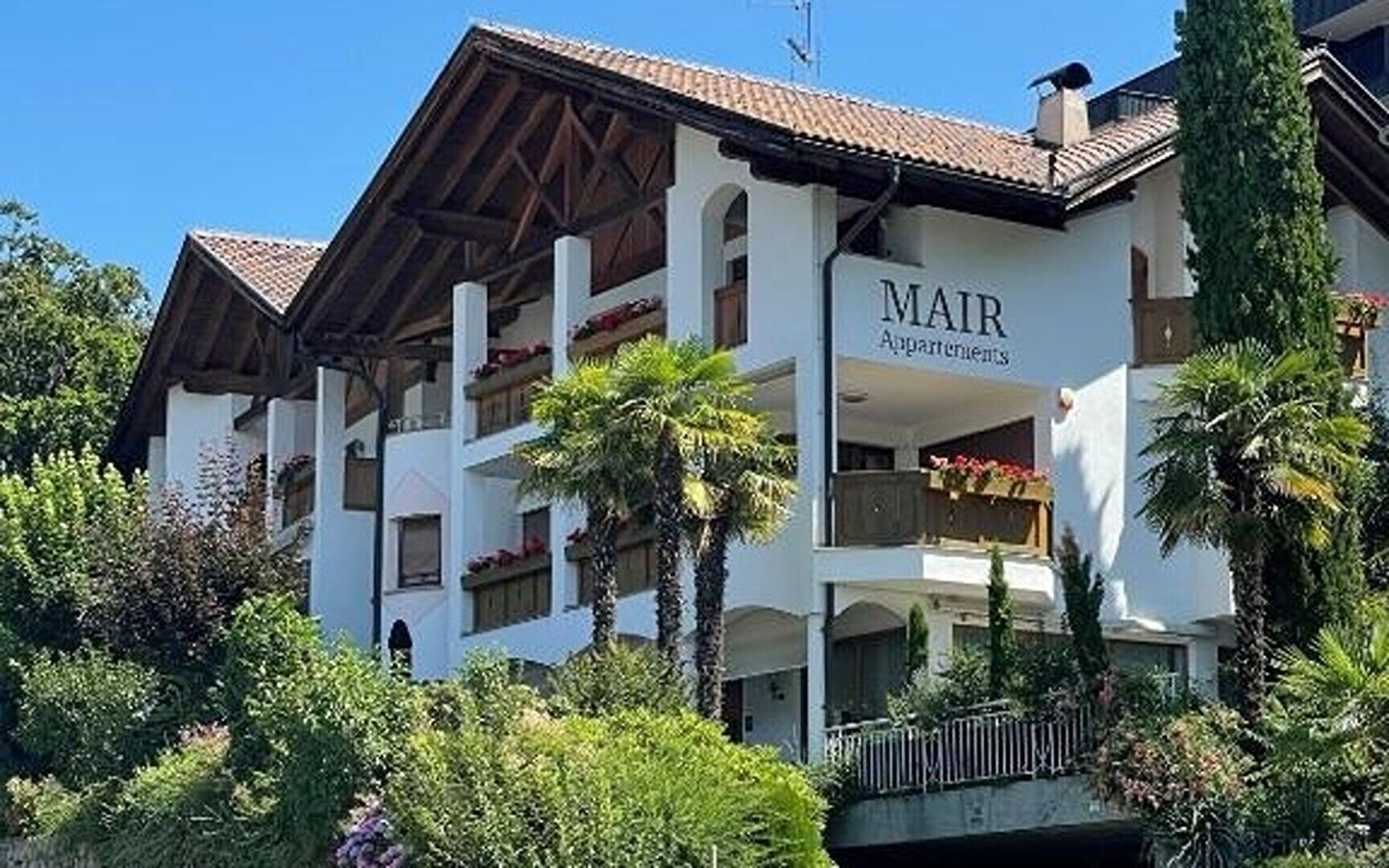 A multi-story building with balconies, labeled 'MAIR Apartments,' surrounded by greenery.