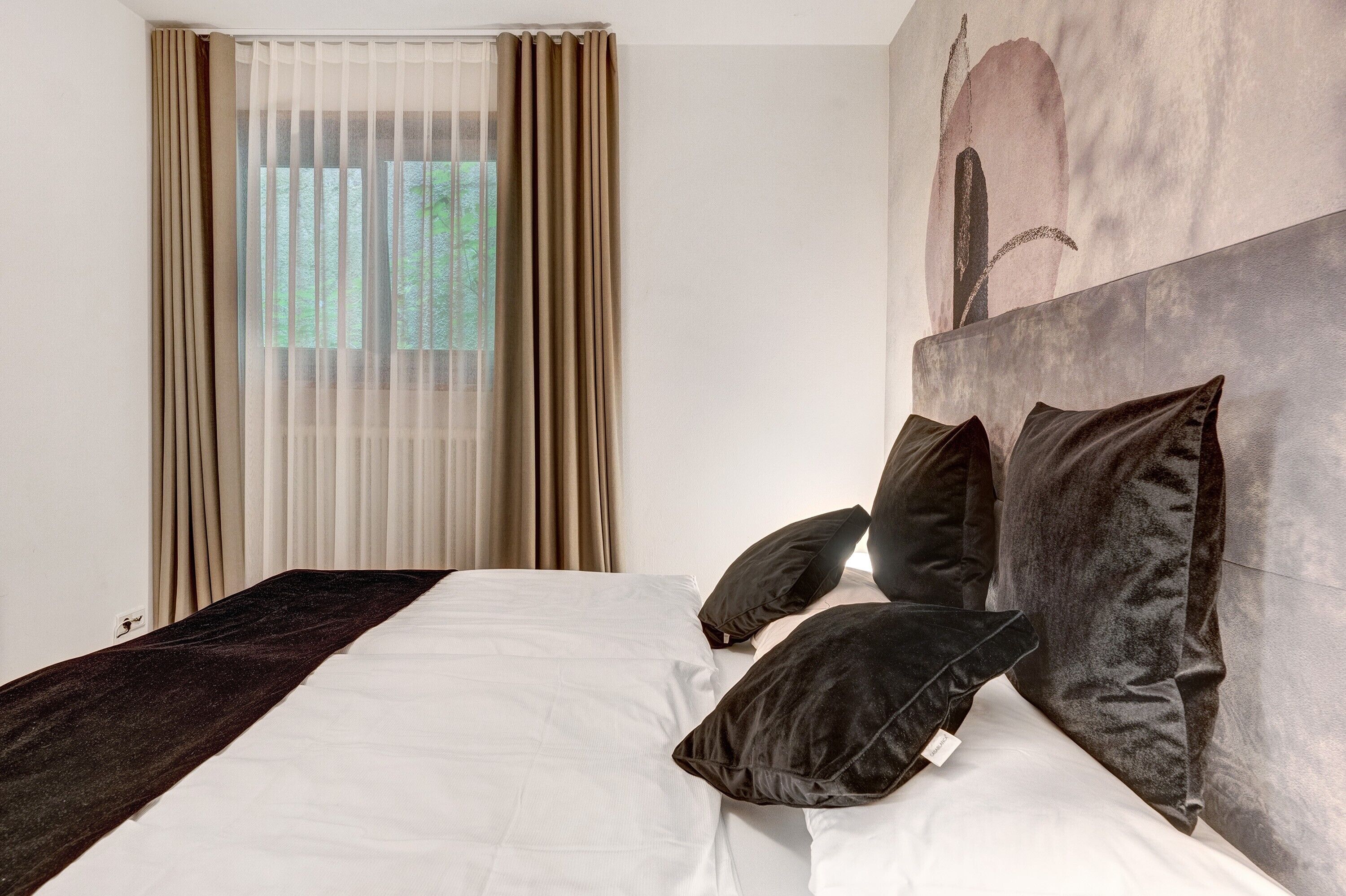 Een hotelkamer met een bed, beige gordijnen, een schilderij aan de muur en uitzicht op groen.