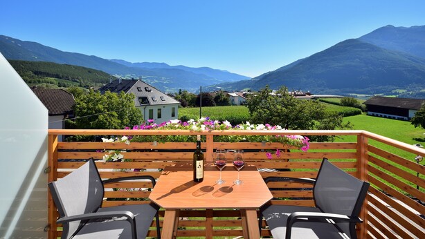 Een houten terras met een tafel en stoelen, met uitzicht op een bergachtig landschap met huizen en begroeiing.