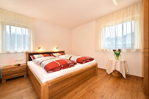 Un dormitorio con una cama de madera, mesitas de noche, una pequeña mesa con un jarrón y ventanas amplias con cortinas translúcidas.