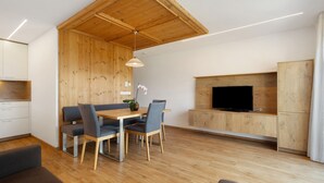Een modern interieur met een houten eettafel, stoelen, een aan een houten wand bevestigde televisie en een bank.