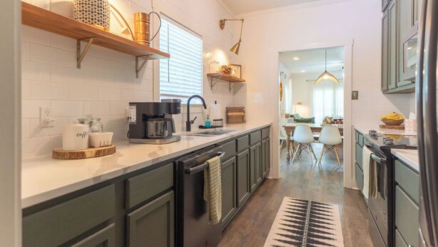 Eine moderne Küche mit dunkelgrünen Schränken, einer weißen Arbeitsplatte und einem schwarzen Kühlschrank. Es gibt eine Kaffeemaschine, einen Wasserkocher und eine Spüle. Die Küche geht in einen Essbereich mit Tisch und Stühlen über.